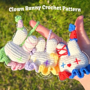 May include: Three handmade crochet clown bunny ornaments in a hand. Each bunny has a white body, colorful accents, and a clown-like design. The text "Clown Bunny Crochet Pattern" is at the top.
