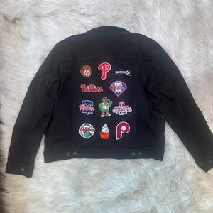 May include: Black denim jacket with multiple Philadelphia Phillies patches. Patches include the Phillies logo, baseballs, and other team-related designs. The jacket has a button-up front and a classic denim jacket style.