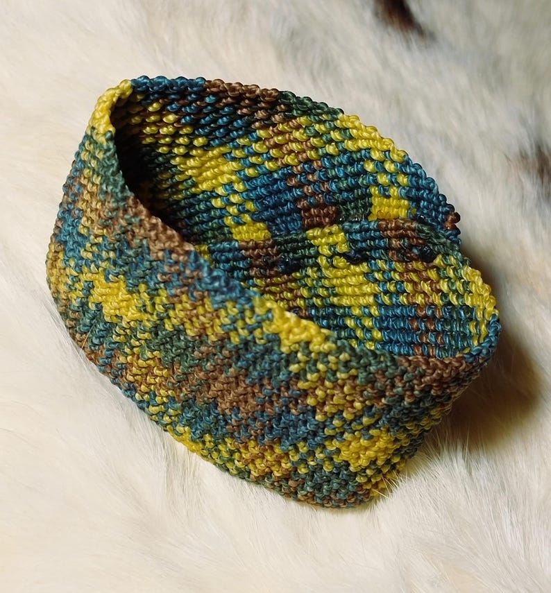K&ouml;nnte beinhalten: Ein kleiner, handgestrickter Korb in Gelb-, T&uuml;rkis- und Braunt&ouml;nen. Das Garn ist eng gewebt und erzeugt eine strukturierte Oberfl&auml;che. Das offene Design des Korbes deutet darauf hin, dass er zur Aufbewahrung oder Dekoration verwendet werden k&ouml;nnte.