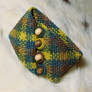 K&ouml;nnte beinhalten: Handgefertigte H&auml;keltasche in Blau-, Gelb- und Braunt&ouml;nen. Die Tasche hat einen Knopfverschluss mit drei Holzkn&ouml;pfen. Das strukturierte Muster und die Farbpalette verleihen ihr einen einzigartigen, handwerklichen Look. Ein stilvolles Accessoire.