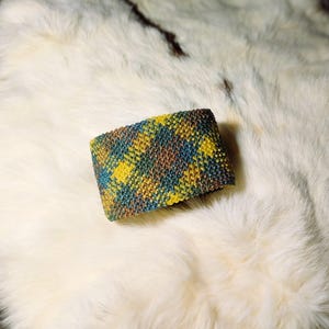 K&ouml;nnte beinhalten: Ein farbenfrohes, gestricktes Armband mit Karomuster. Das Armband zeigt eine Mischung aus gelben, blauen, braunen und gr&uuml;nen F&auml;den. Das Accessoire liegt auf einer wei&szlig;en, strukturierten Oberfl&auml;che, etwa 7 cm breit.