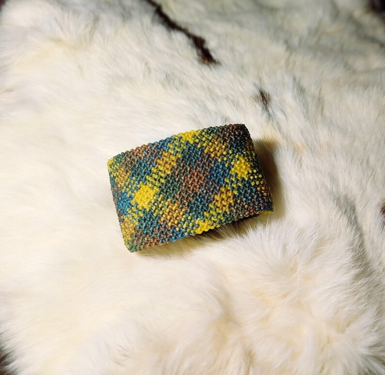 K&ouml;nnte beinhalten: Ein farbenfrohes, gestricktes Armband mit Karomuster. Das Armband zeigt eine Mischung aus gelben, blauen, braunen und gr&uuml;nen F&auml;den. Das Accessoire liegt auf einer wei&szlig;en, strukturierten Oberfl&auml;che, etwa 7 cm breit.