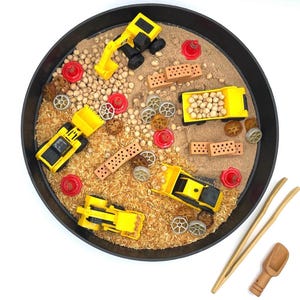 May include: A round black sensory tray filled with light brown sand and wood shavings, containing yellow construction vehicles, small red traffic cones, brown rectangular toy bricks, dried chickpeas, and wheel-shaped pasta pieces, with wooden tongs and a scoop.