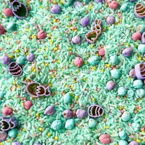 May include: A sensory bin filled with dyed green rice, pastel beads, and Easter shapes. The shapes are eggs and bunnies in purple, pink, and teal. A fun and engaging activity for children, perfect for Easter.
