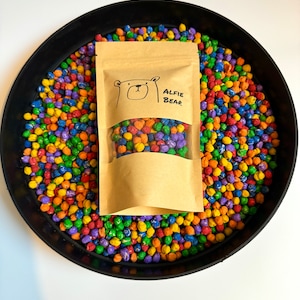 May include: A brown paper bag with a white bear illustration and the text "Alfie Bear" on it. The bag is filled with colourful, small, round objects. The bag is sitting on a black plate filled with the same colourful objects.