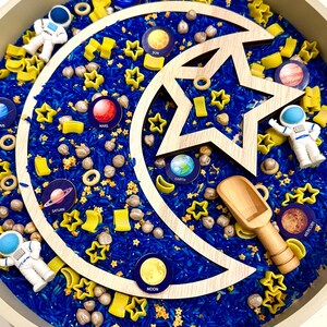 May include: A sensory bin featuring blue dyed rice, wooden star and crescent shapes, yellow star and moon-shaped pasta, and planet and astronaut figurines. Planets include Earth, Mars, Saturn, and Mercury. A wooden scoop is included.