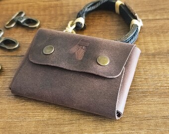 May include: A brown leather wallet with a flap closure secured by two brass-colored snap buttons. The wallet has an embossed design and is next to a braided bracelet and metal clasps.