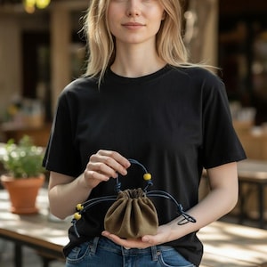 May include: A small olive green drawstring pouch featuring a dark blue cord handle and closure system. The cord is accented with bright yellow spherical beads at the ends and along the handle, creating a functional and compact accessory.