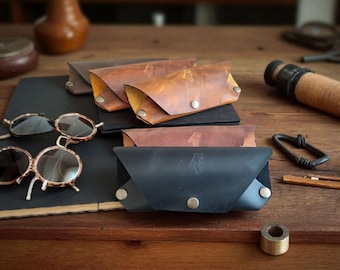 May include: Several leather spectacle cases in black, tan, and brown colours, each featuring a triangular flap secured with a metal snap button. A small embossed logo is visible on the front of each case. Two pairs of glasses are placed nearby.