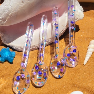 May include: Four clear plastic spoons with a variety of purple, blue, and white glitter and beads embedded in the plastic. The spoons are arranged on a brown surface.