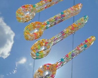 May include: Four clear resin spoons arranged diagonally, each embedded with a variety of colorful, multi-shaped sprinkles including small stars, spheres, and rods in shades of pink, yellow, green, blue, orange, and purple throughout the handles and bowls.