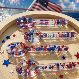 May include: Four clear resin spoons filled with red, white, and blue star-shaped and cylindrical sprinkles. The spoons are arranged on a light-colored wooden surface, accompanied by several loose red, white, and blue star-shaped confetti pieces.