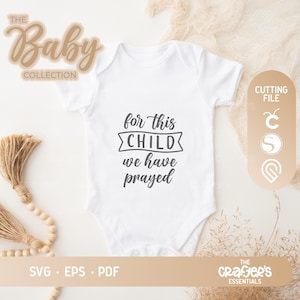 May include: White baby bodysuit with the text "for this child we have prayed" in black lettering. The bodysuit is on a light brown surface with a brown tassel garland.