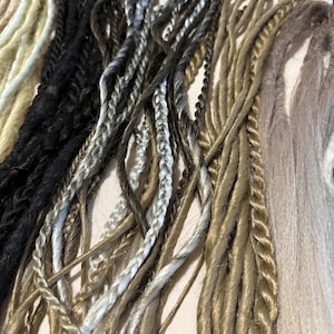 May include: Close-up of several dreadlock extensions in various colors. The colors include blonde, black, gray, and brown. The dreadlocks are made of synthetic hair and are twisted and braided.