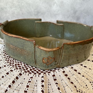 May include: An oval-shaped ceramic bowl with a mottled sage green glaze and a smooth, light-colored interior. The exterior features horizontal ribbed detailing, a small embossed leaf-like emblem, and a brown-toned rim finish.