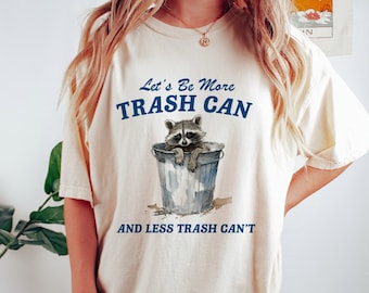 Puede incluir: Una camiseta de color crema con un gráfico azul de un mapache en un contenedor de basura. El texto en la camiseta dice "Let's Be More Trash Can and Less Trash Can't".