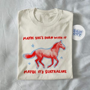 May include: Off-white t-shirt featuring a red horse design with a blue outline and the text "Maybe She's Born With It, Maybe It's Sertraline." Blue stars surround the horse. A "Silly City" sticker is also visible.