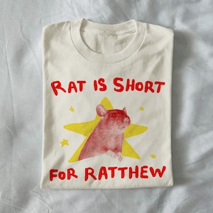 May include: Cream-colored t-shirt with a red rat illustration inside a yellow star. The text "RAT IS SHORT FOR RATTHEW" is printed in red, above and below the graphic. The shirt is folded.