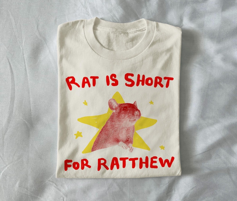 May include: Cream-colored t-shirt with a red rat illustration inside a yellow star. The text "RAT IS SHORT FOR RATTHEW" is printed in red, above and below the graphic. The shirt is folded.