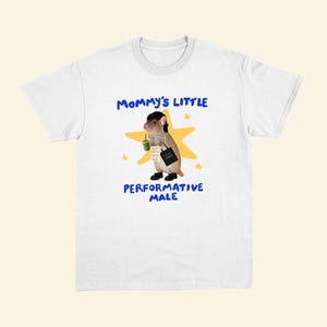 May include: White t-shirt with a graphic of a rodent wearing a black wig, holding a green drink and a tote bag. The text on the shirt reads "Mommy's Little Performative Male" in blue lettering with a yellow star and small stars.