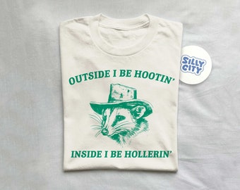 May include: A white t-shirt with a green graphic of a raccoon wearing a cowboy hat. The text "OUTSIDE I BE HOOTIN'" is above the raccoon and the text "INSIDE I BE HOLLERIN'" is below the raccoon.