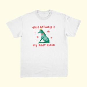 May include: White t-shirt with a graphic of a turquoise horse wearing a yellow hat and red collar. The text "THIS ACTUALLY IS" and "MY FIRST RODEO" are in red, with red star accents.