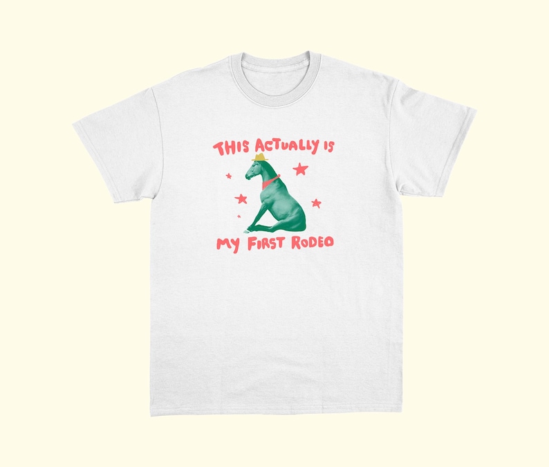 May include: White t-shirt with a graphic of a turquoise horse wearing a yellow hat and red collar. The text "THIS ACTUALLY IS" and "MY FIRST RODEO" are in red, with red star accents.