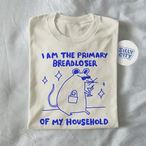 May include: Cream-colored t-shirt with a blue cartoon illustration of a mouse wearing sunglasses and holding a cocktail. The text "I AM THE PRIMARY BREADLOSER OF MY HOUSEHOLD" is printed in blue. A "Silly City" sticker is visible.