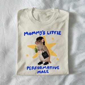 Puede incluir: Camiseta color crema con el texto azul "Mommy's Little Performative Male". La camiseta presenta una ilustración de dibujos animados de un roedor con auriculares, sosteniendo una bebida y una bolsa de mano. El roedor está frente a una estrella amarilla.