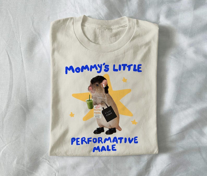 May include: Cream-colored t-shirt with the text "Mommy's Little Performative Male" in blue. The shirt features a cartoon illustration of a rodent wearing headphones, holding a drink, and a tote bag. The rodent is standing in front of a yellow star.