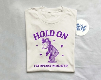 Puede incluir: Una camiseta blanca con texto morado que dice "HOLD ON I'M OVERSTIMULATED". Un oso de dibujos animados morado está en el centro de la camiseta. El oso está de pie sobre sus patas traseras y tiene las patas en el aire. Hay pequeñas estrellas moradas alrededor del oso.