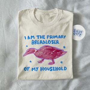 May include: Cream-coloured t-shirt with the text "I AM THE PRIMARY BREADLOSER OF MY HOUSEHOLD" in blue. A pink duck graphic is centered below the text, with blue star accents. A "Silly City" sticker is on the right.