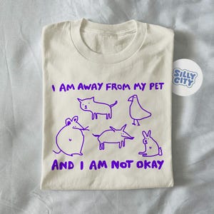 Peut inclure: T-shirt crème avec un dessin violet d'animaux et le texte "I AM AWAY FROM MY PET AND I AM NOT OKAY." Un autocollant "Silly City" est également visible. Le t-shirt est plié.