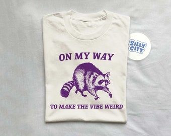 Op de afbeelding: Een wit T-shirt met paarse tekst die "On my way to make the vibe weird" luidt. Boven de tekst staat een paarse illustratie van een wasbeer. Een blauwe en witte sticker met de tekst "Silly City" bevindt zich in de rechterbovenhoek van de afbeelding.