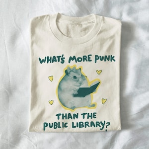 Op de afbeelding: Crèmekleurig T-shirt met een cartoon hamster met een bril en een boek lezend. De tekst luidt "WHAT'S MORE PUNK THAN THE PUBLIC LIBRARY?" met gele hartaccenten. Het shirt is opgevouwen.