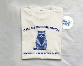 May include: A white t-shirt with blue text that reads "Call me biodegradable because I break down easily". A blue and gray raccoon illustration is centered on the shirt. A blue and white sticker with the text "Silly City" is in the top right corner of the image.