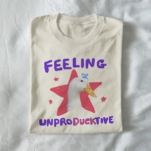 以下が含まれることがあります： 「FEELING UNPRODUCTIVE」の紫色の文字が入ったクリーム色のTシャツ。赤い星の中に漫画のアヒルがいます。アヒルは黄色いくちばしと青いリボンをしています。小さな赤い星がメインのデザインの周りに散らばっています。