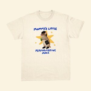 May include: Cream-colored t-shirt with the text "Mommy's Little Performative Male" in blue. The graphic features a cartoon rodent holding a green drink and a black bag, set against a yellow star background. The shirt has a crew neck.