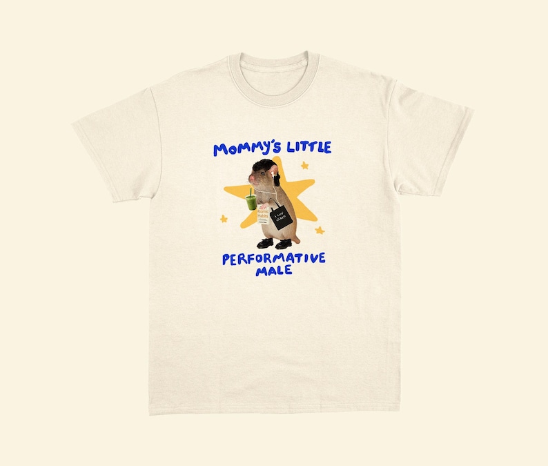 May include: Cream-colored t-shirt with the text "Mommy's Little Performative Male" in blue. The graphic features a cartoon rodent holding a green drink and a black bag, set against a yellow star background. The shirt has a crew neck.