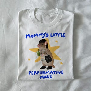 May include: White t-shirt with a graphic of a rodent wearing headphones, holding a green drink and a tote bag. Text on the shirt reads "Mommy's Little Performative Male" in blue. A yellow star is behind the rodent.