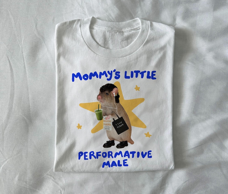 May include: White t-shirt with a graphic of a rodent wearing headphones, holding a green drink and a tote bag. Text on the shirt reads "Mommy's Little Performative Male" in blue. A yellow star is behind the rodent.