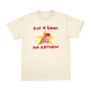 May include: Cream-colored t-shirt with a graphic featuring a red rat inside a yellow star. The text "RAT IS SHORT FOR RATTHEW" is written in red above and below the graphic. The shirt has a crew neck and short sleeves.