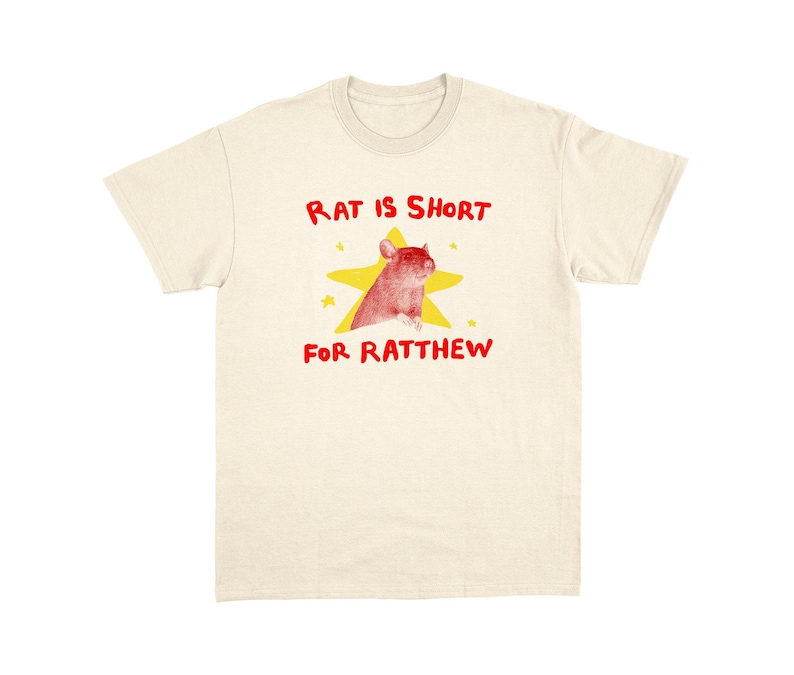 May include: Cream-colored t-shirt with a graphic featuring a red rat inside a yellow star. The text "RAT IS SHORT FOR RATTHEW" is written in red above and below the graphic. The shirt has a crew neck and short sleeves.