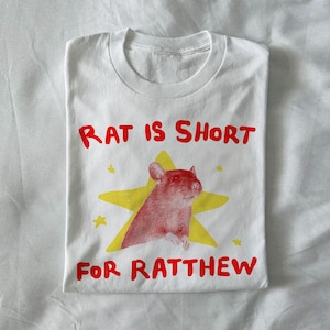 May include: White t-shirt with a red and yellow graphic. The graphic features a red rat inside a yellow star, with the text "RAT IS SHORT FOR RATTHEW" in red letters. The shirt is folded.