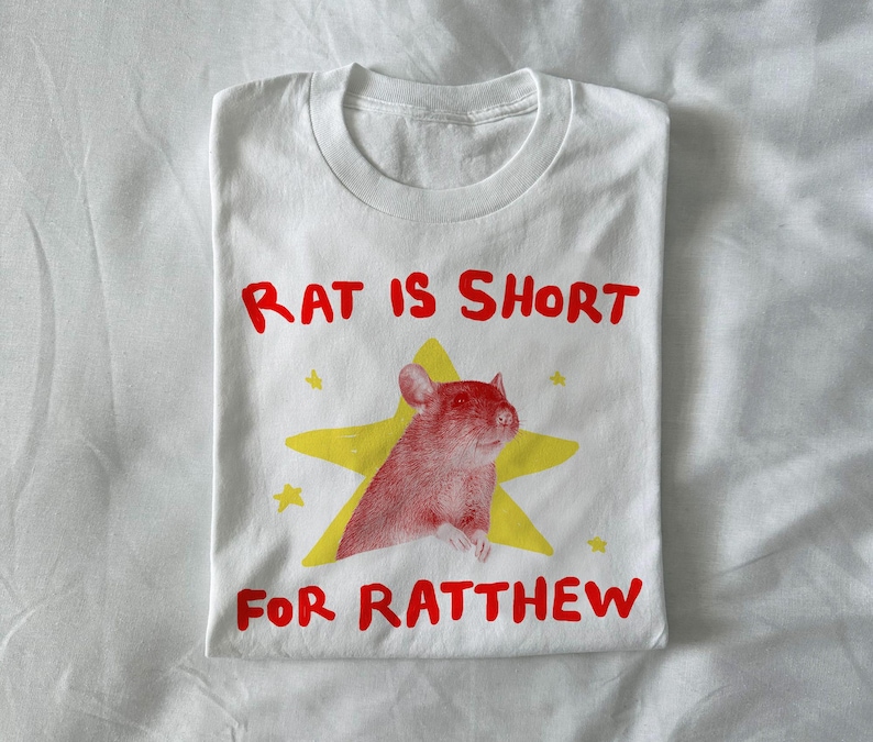 May include: White t-shirt with a red and yellow graphic. The graphic features a red rat inside a yellow star, with the text "RAT IS SHORT FOR RATTHEW" in red letters. The shirt is folded.