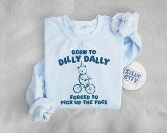 May include: Light blue sweatshirt with a graphic of a bear riding a bicycle. The text on the sweatshirt reads "Born to Dilly Dally, Forced to Pick Up the Pace".