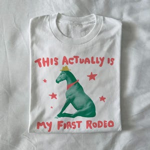 May include: White t-shirt with a graphic of a green horse wearing a cowboy hat and red neckerchief. The text "THIS ACTUALLY IS MY FIRST RODEO" is printed above and below the horse in red.