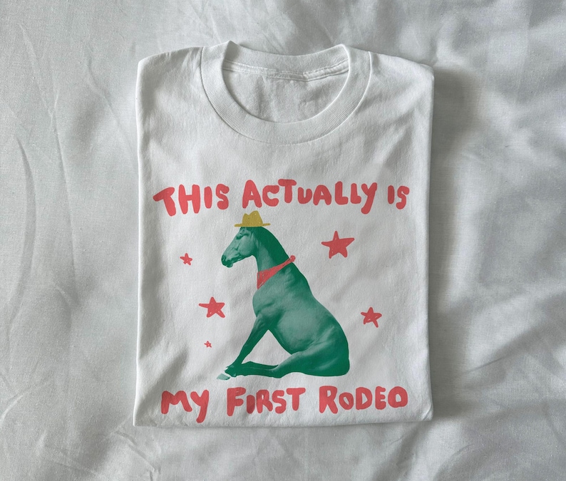 May include: White t-shirt with a graphic of a green horse wearing a cowboy hat and red neckerchief. The text "THIS ACTUALLY IS MY FIRST RODEO" is printed above and below the horse in red.