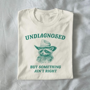 Könnte beinhalten: Gefaltetes cremefarbenes T-Shirt mit grünem Text "UNDIAGNOSED" und "BUT SOMETHING AIN'T RIGHT". Darunter eine grüne Illustration eines Waschbären mit Cowboyhut.
