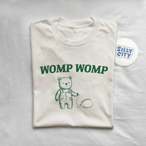 Può includere: Una t-shirt color crema con una grafica verde di un orso che tiene un palloncino. Il testo "WOMP WOMP" è stampato in verde sopra l'orso. Un adesivo blu e bianco con il testo "SILLY CITY" si trova nell'angolo in alto a destra dell'immagine.
